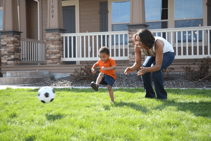 The image shows a woman and young boy playing soccer together in a grassy yard in front of a house.